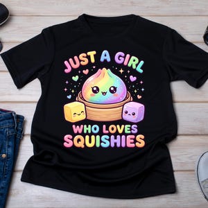 May include: A black short-sleeve t-shirt featuring a colorful graphic of a rainbow-colored dumpling in a bamboo steamer, flanked by a yellow and a purple cube. The text reads JUST A GIRL WHO LOVES SQUISHIES in pastel letters with small hearts.