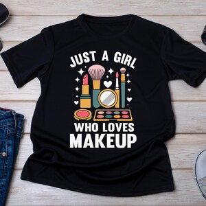 Pode incluir: Camiseta preta com a frase "JUST A GIRL WHO LOVES MAKEUP" e gráficos temáticos de maquiagem. O design inclui batom, um pincel de maquiagem, um espelho compacto e uma paleta de sombras. A camiseta é cercada por acessórios como óculos de sol, jeans e tênis.