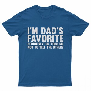 May include: A blue t-shirt with the text "I'M DAD'S FAVORITE" in large white letters. Below, smaller text reads "SERIOUSLY, HE TOLD ME NOT TO TELL THE OTHERS". The shirt is a casual wear item.