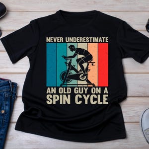 Op de afbeelding: Zwart t-shirt met een retro ontwerp van een fietser op een spinningfiets. De tekst luidt "NEVER UNDERESTIMATE AN OLD GUY ON A SPIN CYCLE" in het wit. Zonnebril, horloge en jeans zijn ook zichtbaar.