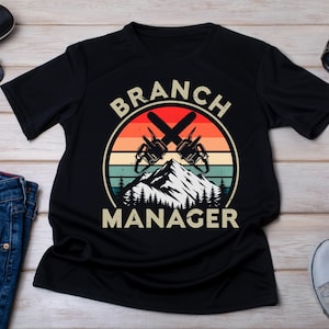 Puede incluir: Camiseta negra con un gráfico de estilo vintage de una cordillera y dos motosierras cruzadas. El texto "BRANCH MANAGER" está impreso debajo del gráfico.