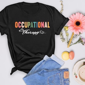 May include: Black t-shirt with the words "OCCUPATIONAL Therapy" in colorful, playful lettering. The shirt is paired with blue jeans, a pink flower, and a cup of coffee, creating a casual, inviting aesthetic.