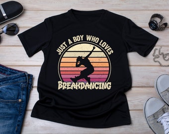 Puede incluir: Camiseta negra con un gráfico retro de una persona bailando breakdance. El gráfico está rodeado de un círculo con el texto "Just a boy who loves breakdancing".