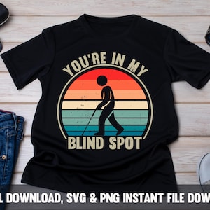 May include: Black t-shirt with a retro design featuring a stick figure with a cane walking in front of a sunset. The text "YOU'RE IN MY BLIND SPOT" is arched above and below the figure. Sunglasses, a watch, and jeans are in the background.