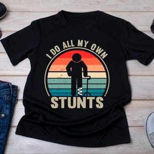 May include: Black t-shirt featuring a vintage-style graphic of a person with a broken leg and a walking stick. The graphic reads "I do all my own stunts".