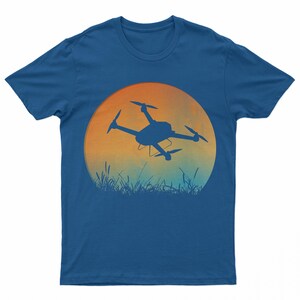May include: A blue t-shirt with a graphic of a drone flying in front of a sunset. The drone is a silhouette against the orange and blue sky.