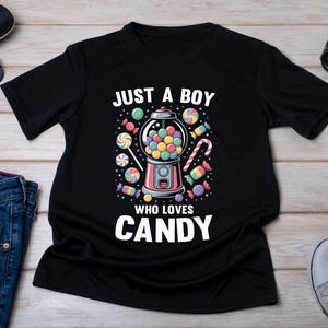 Może przedstawiać: Czarny t-shirt z krótkim rękawem z grafiką automatu do gum do żucia otoczonego różnymi kolorowymi słodyczami. Tekst nad automatem brzmi JUST A BOY, a tekst poniżej brzmi WHO LOVES CANDY, zapisany pogrubionymi białymi wielkimi literami.