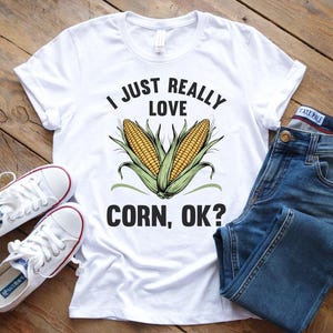 Op de afbeelding: Een wit t-shirt met een afbeelding van twee gele maïskolven met groene bladeren en de zwarte tekst I JUST REALLY LOVE CORN, OK? Het t-shirt wordt getoond naast een paar witte canvas sneakers en een blauwe spijkerbroek.