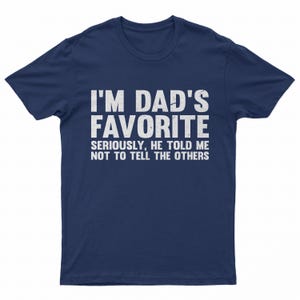 May include: Navy blue t-shirt with white text that reads "I'M DAD'S FAVORITE" in large capital letters. Below, smaller text says "SERIOUSLY, HE TOLD ME NOT TO TELL THE OTHERS". The shirt is a casual wear item.
