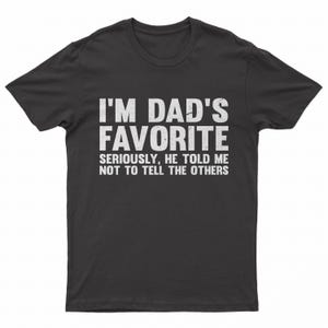 May include: A dark gray t-shirt with the text "I'M DAD'S FAVORITE" in large white letters. Below, smaller text reads "SERIOUSLY, HE TOLD ME NOT TO TELL THE OTHERS."