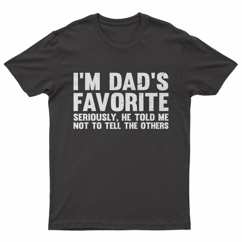 May include: A dark gray t-shirt with the text "I'M DAD'S FAVORITE" in large white letters. Below, smaller text reads "SERIOUSLY, HE TOLD ME NOT TO TELL THE OTHERS."