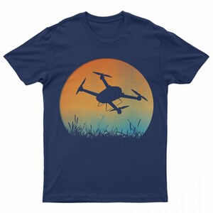 May include: A navy blue t-shirt with a silhouette of a drone flying in front of a sunset. The sunset is a gradient of orange, yellow, and blue.