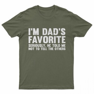 May include: Olive green t-shirt with the text "I'M DAD'S FAVORITE" in large white letters. Below, smaller text reads "SERIOUSLY, HE TOLD ME NOT TO TELL THE OTHERS". A casual, humorous tee.