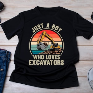 May include: Black t-shirt with a graphic of a yellow excavator on a sunset background. Text reads "JUST A BOY WHO LOVES EXCAVATORS" in white. The shirt is displayed with sunglasses, jeans, and sneakers.