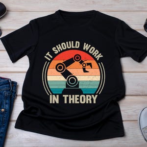May include: Black t-shirt with a retro design featuring a robotic arm and the text "IT SHOULD WORK IN THEORY." The design is set against a striped background of orange, red, and teal. Accessories include sunglasses, jeans, and sneakers.