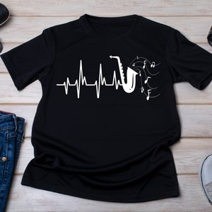 Peut inclure: T-shirt noir avec un graphique blanc d'un saxophone avec des notes de musique et une ligne de battement de cœur. Le texte "My heart beats for jazz" est imprimé sur le t-shirt.