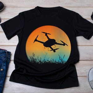 May include: Black t-shirt with a graphic of a drone flying in front of a sunset. The sunset is orange, yellow, and blue.