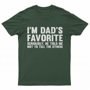 May include: Dark green t-shirt with the text "I'M DAD'S FAVORITE" in large white letters. Below, smaller text reads "SERIOUSLY, HE TOLD ME NOT TO TELL THE OTHERS". A humorous graphic tee.