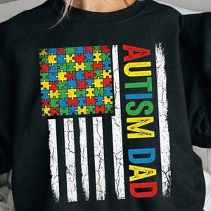 May include: Black sweatshirt featuring a design with a puzzle piece flag and the text "AUTISM DAD" in colorful letters. The flag design includes a square of colorful puzzle pieces and white stripes.