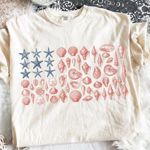 Puede incluir: Una camiseta de manga corta color crema con un diseño gráfico dispuesto en un patrón de bandera rectangular. La sección izquierda muestra estrellas de mar azules, mientras que la derecha presenta varias conchas en tonos coral y rosa.