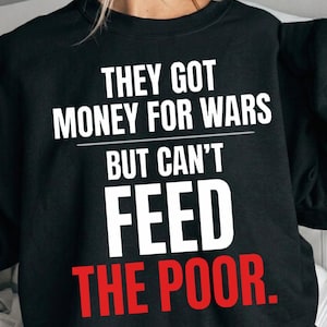 Op de afbeelding: Zwart sweatshirt met witte tekst: "THEY GOT MONEY FOR WARS BUT CAN'T FEED THE POOR." De woorden "FEED THE POOR" zijn rood. Het sweatshirt heeft een ronde hals.