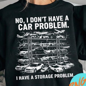 Puede incluir: Una sudadera negra de cuello redondo con un estampado gráfico blanco de una estantería de varios niveles llena de varios coches y un avión. El texto dice: NO, I DON'T HAVE A CAR PROBLEM. I HAVE A STORAGE PROBLEM. El diseño incluye detalles de rayos.