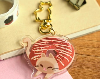 May include: A clear acrylic keychain featuring a cartoon cat wearing a donut with white frosting and red sprinkles. The donut has a hole in the center and the cat is smiling. The keychain has a gold metal ring and a gold metal cat-shaped charm.