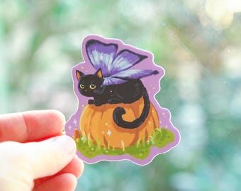 May include: A black cat with purple butterfly wings sits on top of an orange pumpkin. The cat has yellow eyes and a black nose. The pumpkin is surrounded by green grass.