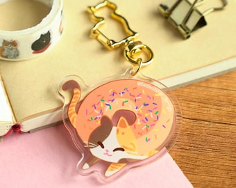 May include: A clear acrylic keychain featuring a cartoon cat curled up inside a pink donut with colorful sprinkles. The keychain has a gold metal ring and clasp.