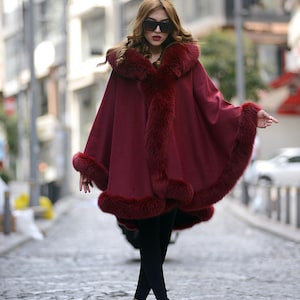 May include: A deep burgundy cape featuring a thick, matching faux fur trim along the hood, front opening, and hemline. The garment has a loose, flowing silhouette that drapes over black leggings and is paired with neutral-toned heeled sandals.