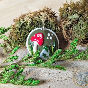 Könnte beinhalten: Ein grüner und weißer Keramik-Anhänger mit einem roten und weißen Pilz, einem kleinen Stein und grünem Laub.