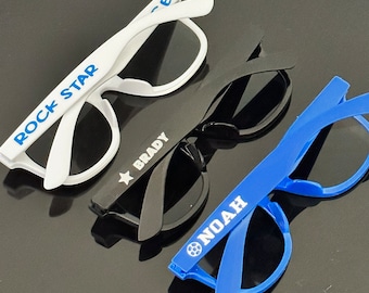 May include: Three pairs of sunglasses with different coloured frames and lenses. The white pair has "ROCK STAR" written on the frame. The black pair has a white star and "BRADY" written on the frame. The blue pair has a football and "NOAH" written on the frame.