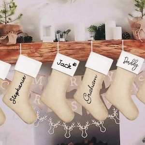 Op de afbeelding: Een rij beige kerstsokken met witte manchetten hangt aan een houten schoorsteenmantel. Elke sok is gepersonaliseerd met een naam in zwart script: Stephanie, Jack, Graham en Daddy. Naast de naam Jack staat een pootafdruk.