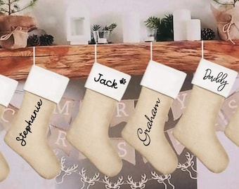 May include: A row of beige Christmas stockings with white cuffs hang from a wooden mantel. Each stocking is personalized with a name in black script: Stephanie, Jack, Graham, and Daddy. A paw print is next to the name Jack.
