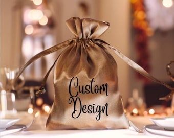 May include: A gold satin drawstring bag with the words "Custom Design" in black script. The bag is gathered at the top with long, thin drawstrings. The background is blurred, with warm, ambient lighting.