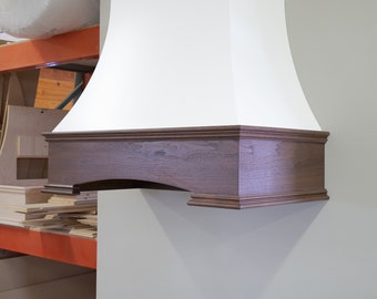 May include: A white kitchen range hood with a brown wood trim. The hood is mounted on a wall and has a curved top.