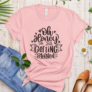 以下が含まれることがあります： 「Oh honey I'm just getting started」と黒で書かれたグラフィックがプリントされたピンクのTシャツです。