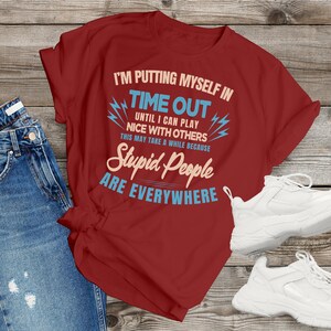 May include: A maroon t-shirt with the text "I'M PUTTING MYSELF IN TIME OUT" and the phrase "Stupid People ARE EVERYWHERE". The shirt is paired with blue jeans and white sneakers.
