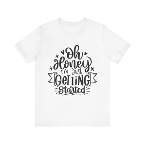 以下が含まれることがあります： 「Oh Honey I'm Just Getting Started」の黒い文字がプリントされた白いTシャツ。デザインにはハート、星、装飾的な要素が含まれています。カジュアルな普段着のTシャツ。