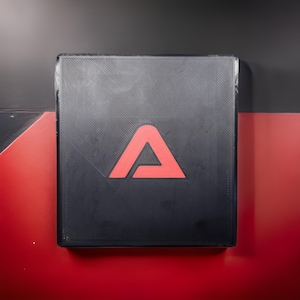 Op de afbeelding: Een zwarte vierkant met een rood driehoekig logo. De vierkant heeft een gestructureerd oppervlak en staat tegen een rode en zwarte achtergrond. Het logo is een gedurfd, eenvoudig ontwerp.