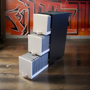 Può includere: Un'unità di stoccaggio nera con tre cassetti bianchi, ciascuno contenente cartucce di gioco grigie. I cassetti hanno maniglie curve. L'unità è su un pavimento di legno, con graffiti sullo sfondo.