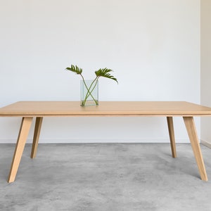 Peut inclure: Une table à manger en bois clair avec un plateau rectangulaire et quatre pieds inclinés. La table est dans une pièce avec un mur blanc et un sol en béton. Un petit vase en verre avec des feuilles vertes est posé sur la table.