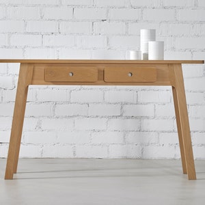 Op de afbeelding: Een lichte houten consoletafel met twee lades. De tafel heeft een eenvoudig, modern ontwerp met schuine poten.