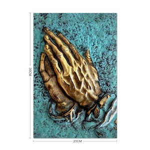 Puede incluir: Una escultura de pared 3D de dos manos juntas en oración. Las manos están hechas de metal dorado y están colocadas sobre un fondo azul turquesa.