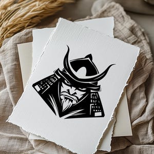 K&ouml;nnte beinhalten: Schwarz-Wei&szlig;-Illustration eines Samurai-Kriegers auf strukturiertem, wei&szlig;em Papier. Der Samurai wird mit Helm und Bart dargestellt. Das Kunstwerk liegt auf einem neutralfarbenen Stoff, was auf einen Druck oder ein Kunstwerk f&uuml;r die Wand hindeutet.