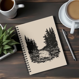 May include: Spiral-bound notebook featuring a black and white illustration of a forest scene with a river. The notebook has a cream-colored cover and a black spiral binding. A pen and two cups of coffee are also visible.