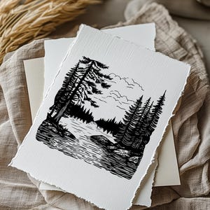 May include: Black and white art print depicting a serene forest landscape with a lake and tall pine trees. The print has a deckled edge and is layered with other white and cream-colored paper. The image is set on a neutral-toned fabric.