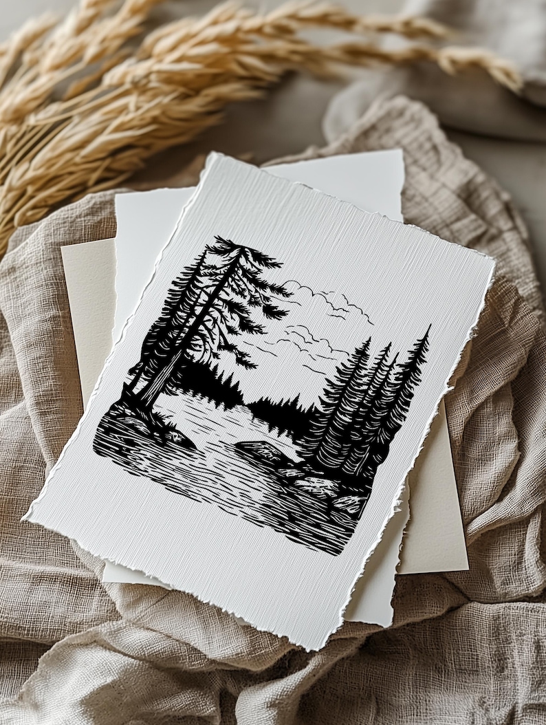 May include: Black and white art print depicting a serene forest landscape with a lake and tall pine trees. The print has a deckled edge and is layered with other white and cream-colored paper. The image is set on a neutral-toned fabric.