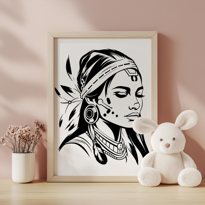 May include: Black and white framed art print of a Native American woman with closed eyes, wearing a detailed headdress and jewelry. The artwork is displayed on a wooden surface with a pink background, alongside a white stuffed animal and a vase of dried flowers.