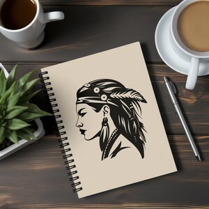 May include: A spiral-bound notebook with a cream-colored cover featuring a black and white illustration of a Native American woman in profile. The woman wears a headband, feather, and earrings. The notebook is on a dark wooden surface, next to a pen and coffee cups.